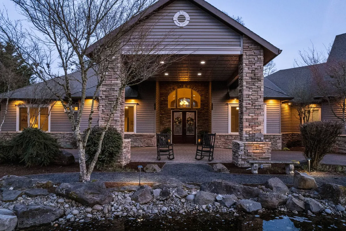 The image features the main entrance to Punkin Manor, with inviting rocking chairs on a porch, a stone facade, lush landscaping, and a tranquil water feature. The welcoming entry hints at the peaceful, retreat-like setting in the heart of wine country.