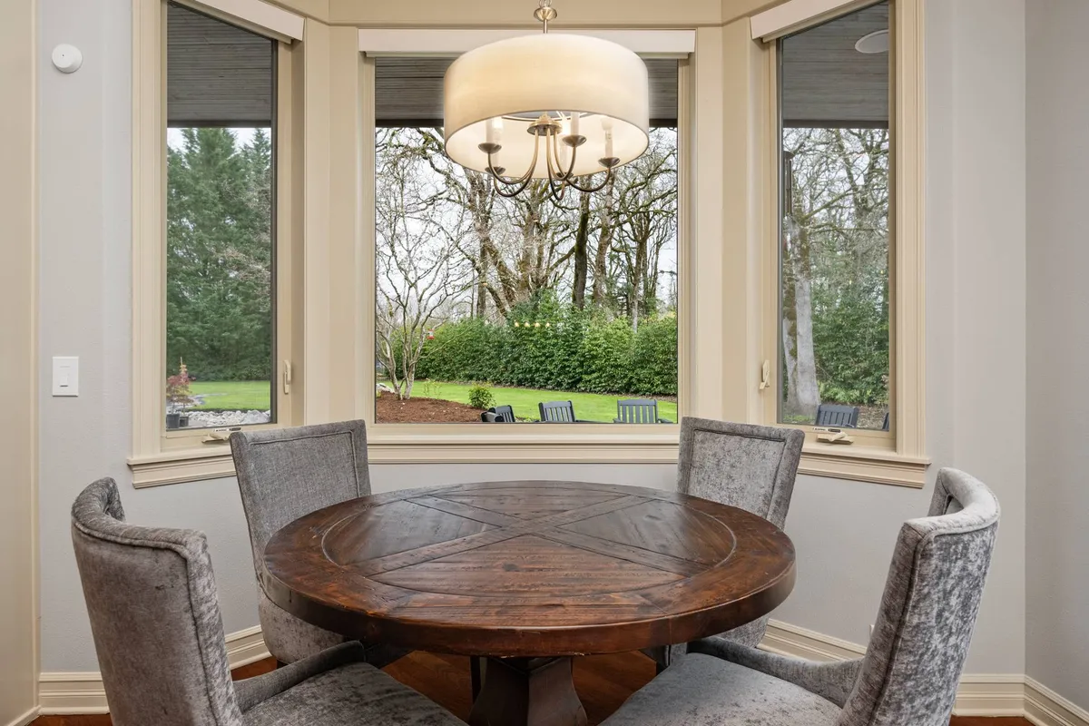 A round wooden table sits beneath a chandelier, framed by large windows that reveal the lush greenery outside. The space is bathed in natural light and offers a peaceful transition between interior comfort and the beckoning outdoors, perfect for planning or unwinding from a day of adventure.