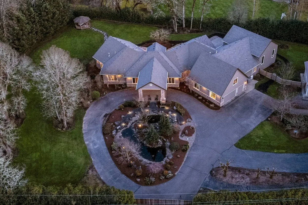The image showcases a sprawling, beautifully designed single-story craftsman estate surrounded by lush, landscaped gardens. A tranquil pond glistens in the foreground, while a wide circular driveway leads to the inviting entrance of the home. The setting is serene and welcoming, ideal for gatherings, celebrations, and quiet outdoor moments.