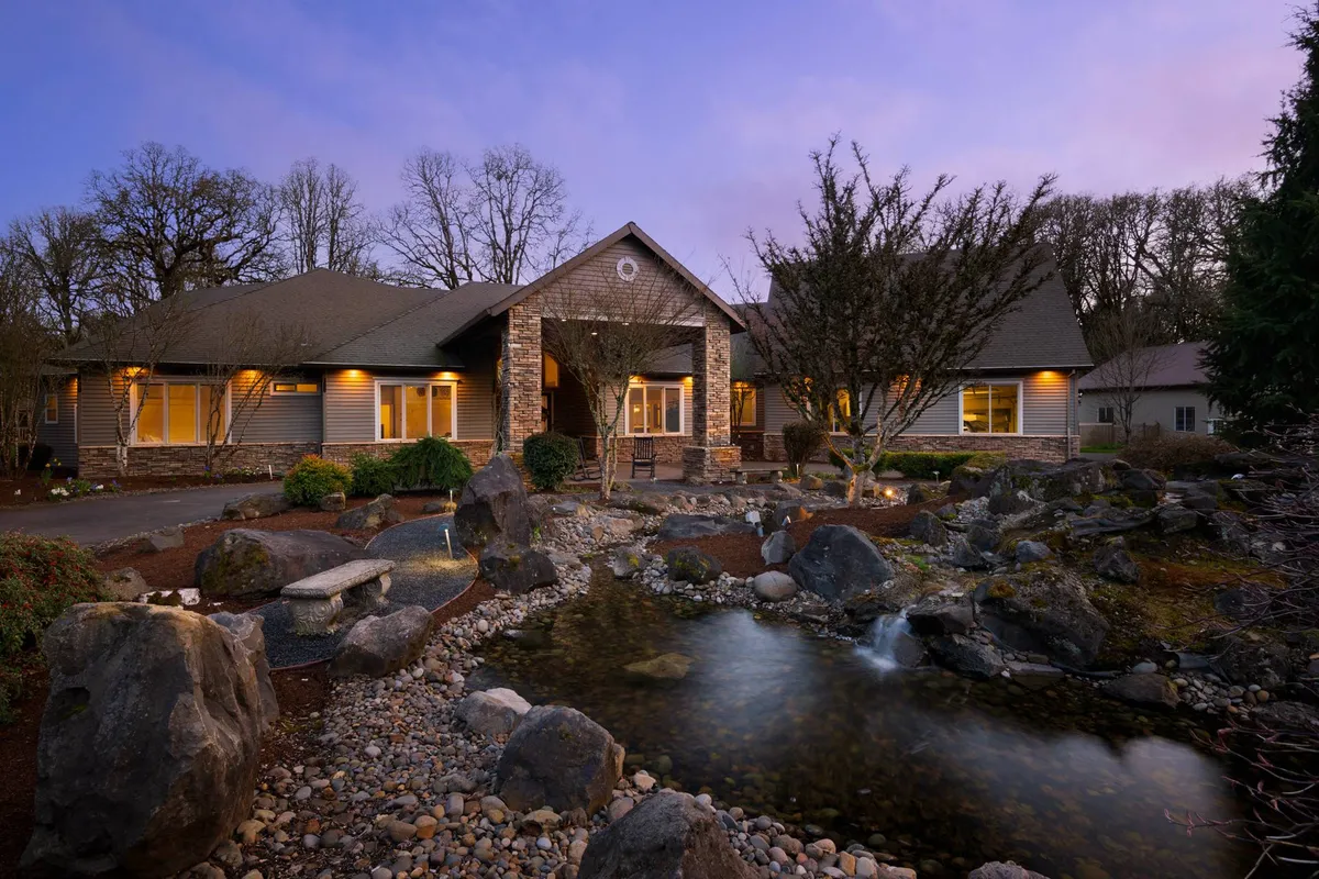 An elegant view of the front entrance features a landscaped garden with a water feature and natural rock formations. The mature trees and curated greenery stretch across a wide, fully fenced area, enhancing the sense of a private retreat.