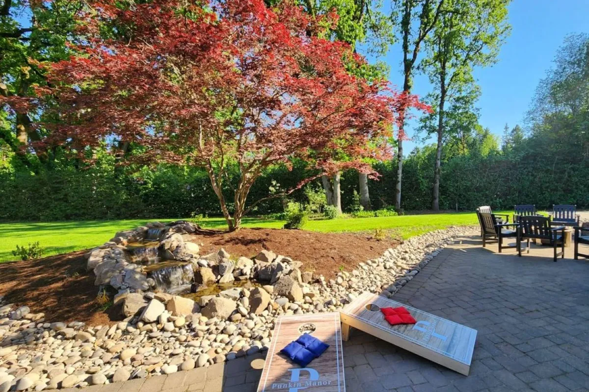 This exterior image features a vibrant red tree shading a small waterfall, a seating area perfect for socializing, and a cornhole game set up on the lawn. The scene radiates a cheerful, leisurely mood ideal for families gathering outdoors, with ample space for children and adults to enjoy the grounds together.