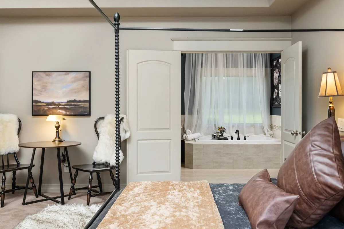 A tranquil, luxurious bedroom with a bed adorned in decorative pillows, a bathtub subtly positioned behind curtains, and calming artwork. A bedside table with a lamp creates a soothing atmosphere, perfect for relaxation and private moments of self-care.