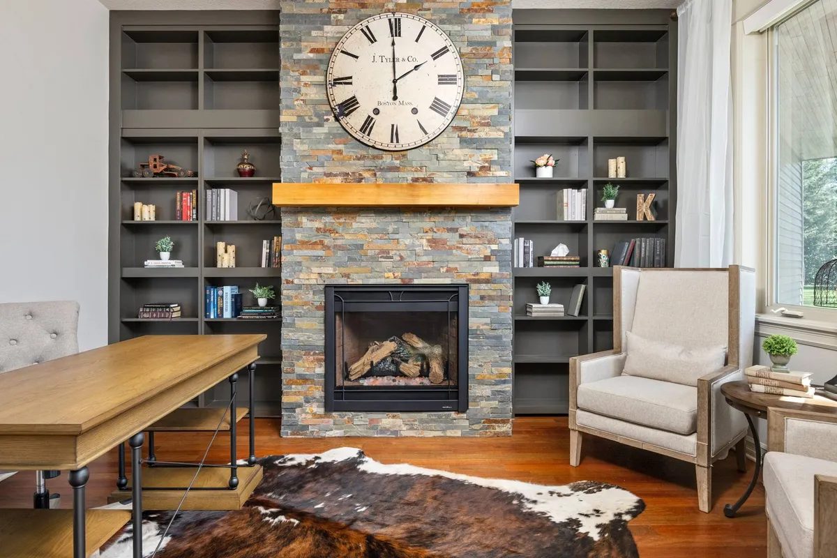 A cozy corner with a stone fireplace, large wall clock, cowhide rug, and bookshelves. There's a desk space suitable for working, as well as a comfortable area for relaxed conversations or creative brainstorming by the fire.