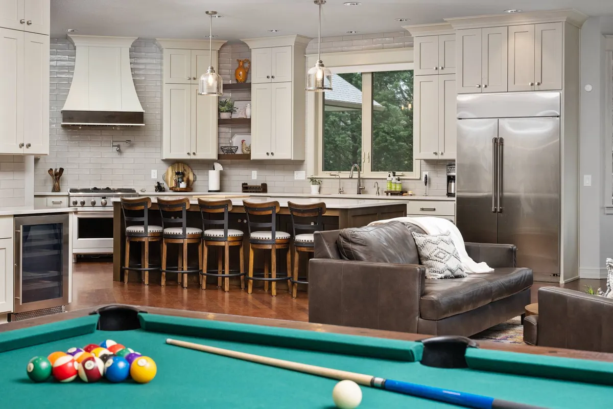 This image displays a lively and welcoming multipurpose room featuring a pool table, kitchen island, and a comfortable sofa set against large windows offering views of the outdoors. The layout suggests ample space for both social gatherings and active recreation, with natural light and a sense of openness that hints at access to the surrounding landscape.