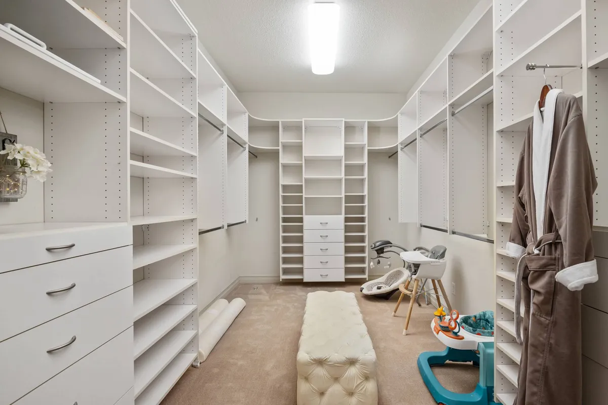 A functional and calm walk-in closet with ample shelving, a plush ottoman, and visible baby gear. Elements such as a robe and organized storage reveal thoughtful amenities for families and make routines easy and peaceful.