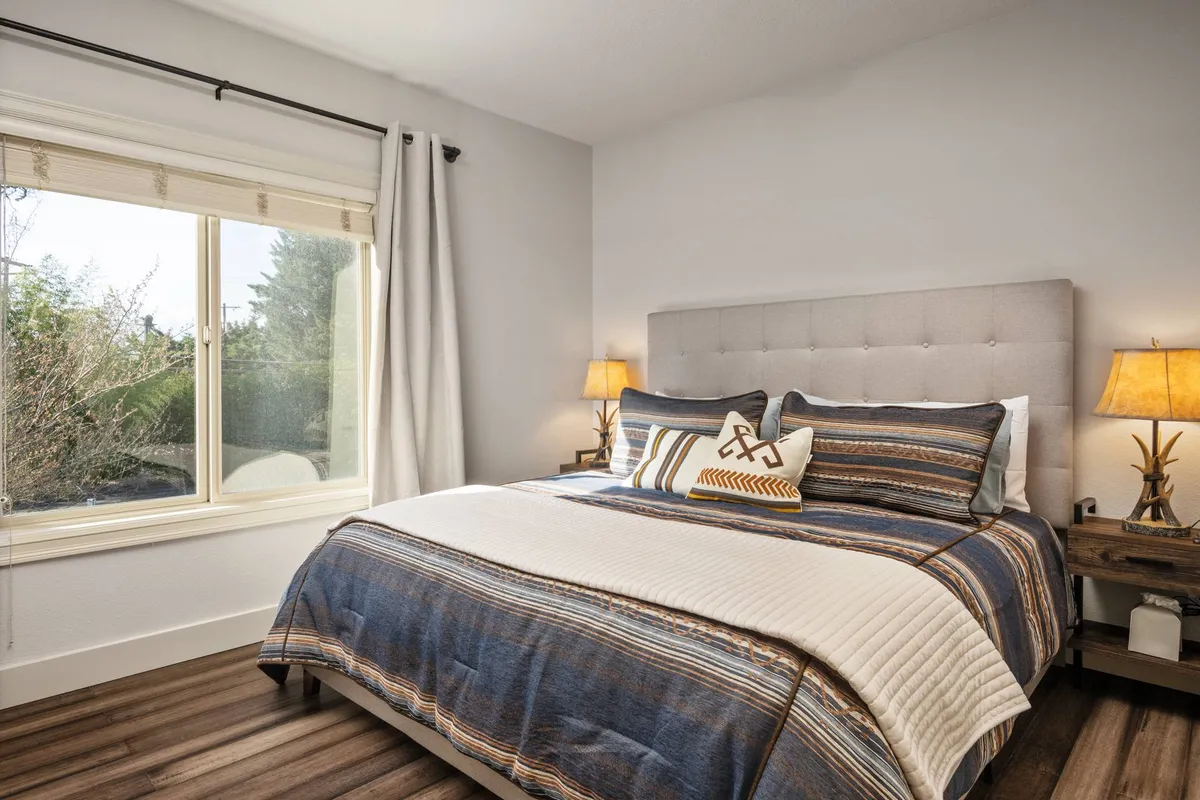 A cozy and serene bedroom featuring a comfortable bed with decorative pillows, illuminated by soft natural light streaming through a large window. Two bedside lamps add a warm ambiance, and the scene invites rest and relaxation, ideal for unwinding at the end of the day.