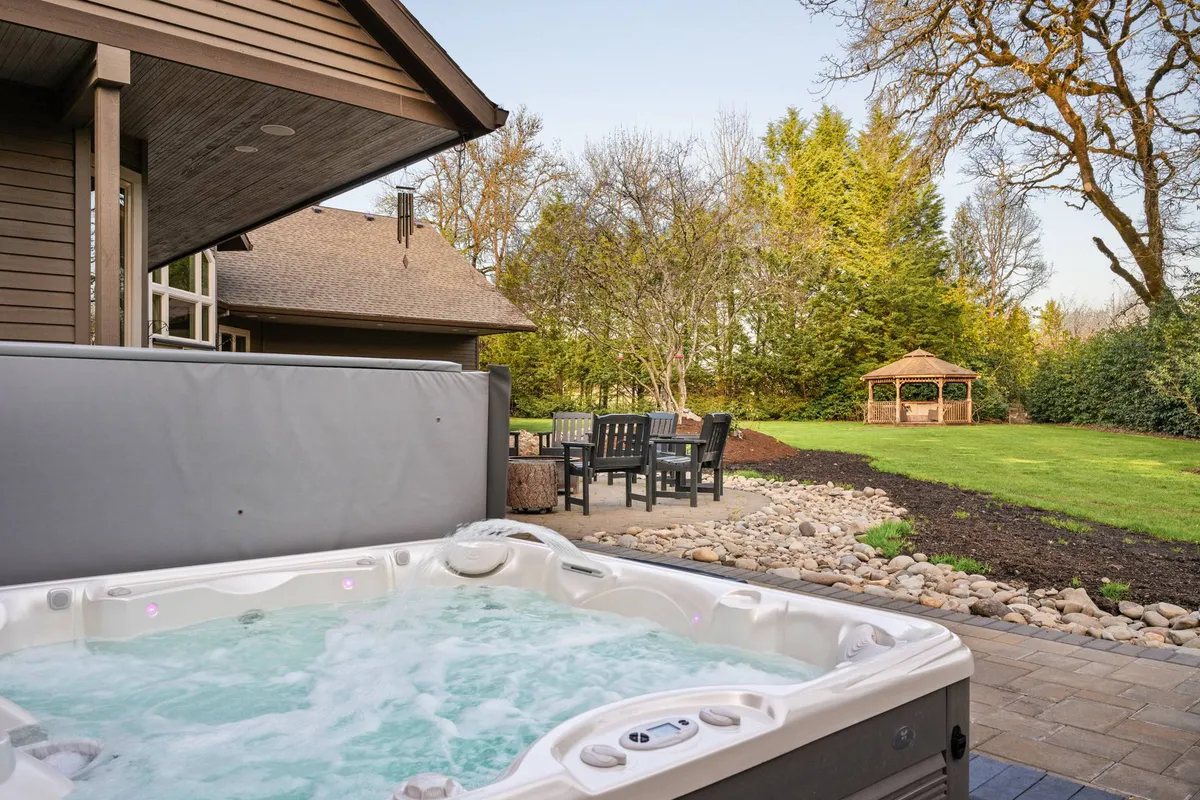 This exterior image captures a tranquil hot tub nestled beside a gazebo, set against a lush backdrop of greenery. Comfortable outdoor seating and mature landscaping create an inviting atmosphere, while the expansive lawn visible beyond hints at space for both relaxation and play.